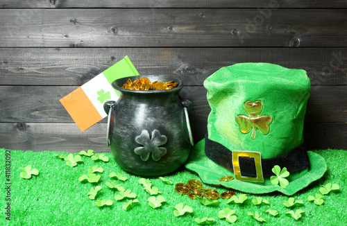 Cast iron pot with treasure, irish flag with clover leaf and leprehaun hat, clover leaves and coins on green grass, dark wooden planks background. Saint Patricks Day banner, poster, flyer, invitation