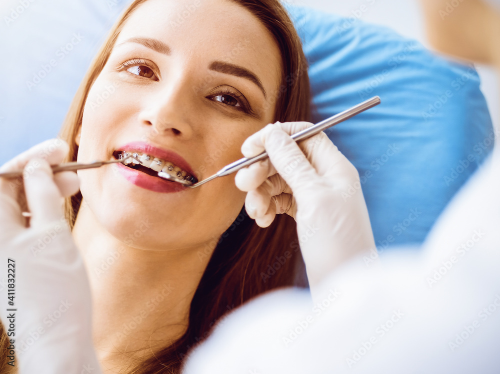 Smiling young woman with orthodontic brackets examined by dentist in sunny dental clinic. Healthy teeth and medicine concept Smiling young woman with orthodontic brackets examined by dentist in sunny dental clinic. Healthy teeth and medicine concept