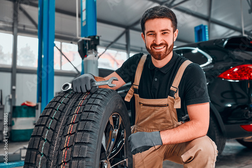 A technician in workwear, holding a wrench and a car tire in tire fitting.