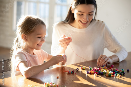 Happy caring young Caucasian mother and little 6s daughter have fun together making bracelets string colorful beads on thread. Smiling mom and small girl child involved in funny hobby activity. photo