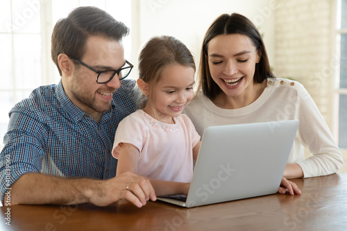 Smiling young Caucasian family with small 7s daughter look at laptop screen watching funny video online. Happy mom and dad have fun using computer with little girl child at home. Technology concept.