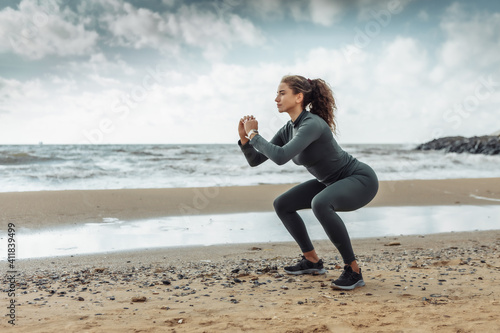 Curly sports woman in sportswear doing squats exercise on beach. Healthy lifestyle