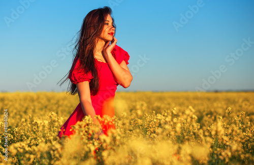 Beautiful young girl in a red dress posing in a field with canola. Happy woman on the nature. Spring season, warm day.