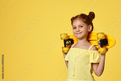 happy little child girl in t-shirt holding yellow longboard on shoulders over yellow background