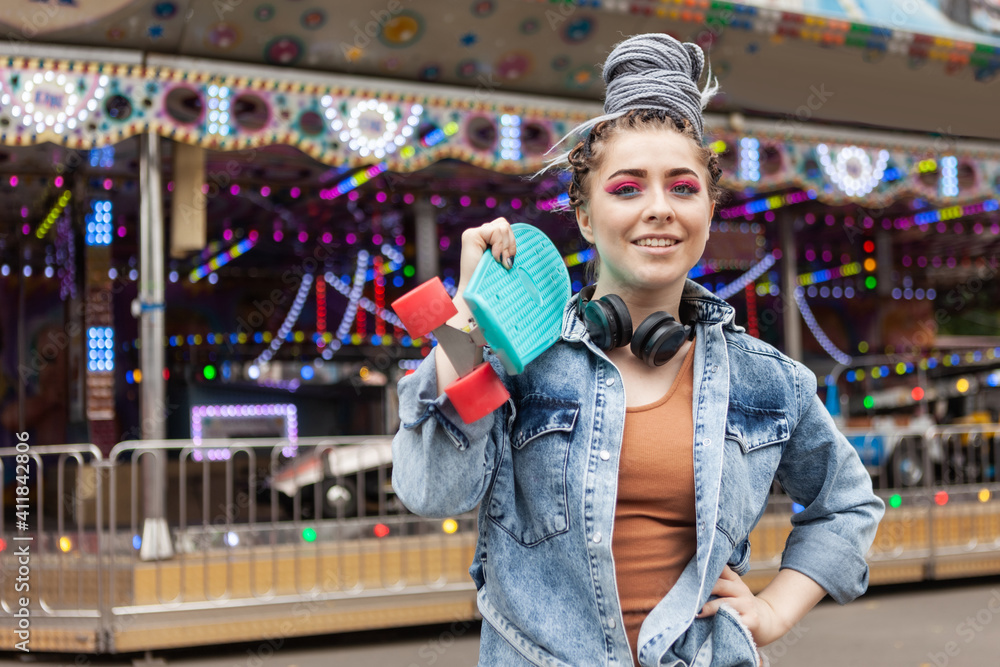 lifestyle portrait of an extraordinary hipster woman with african braids dressed in a denim jacket with a skateboard in an amusement park