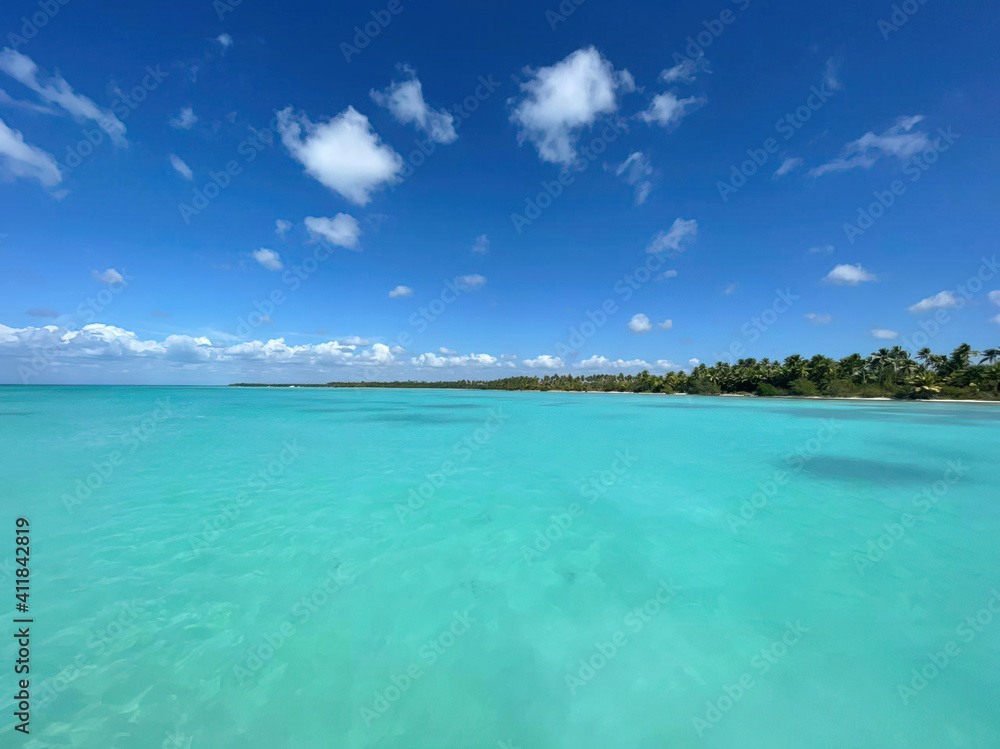 Ocean or sea calm water surface under a blue sky with a few clouds. View on the shore with a beach and tropical trees. Vacation resort paradise in summer time