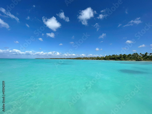 Fototapeta Naklejka Na Ścianę i Meble -  Ocean or sea calm water surface under a blue sky with a few clouds. View on the shore with a beach and tropical trees. Vacation resort paradise in summer time