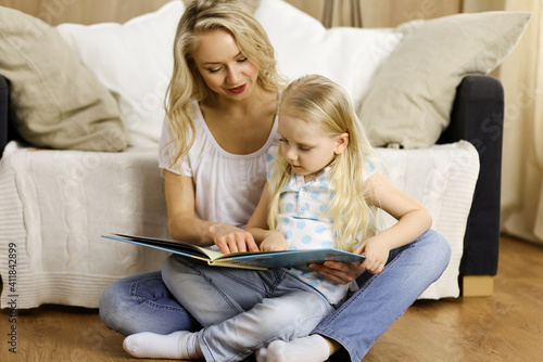 Happy family. Blonde young mother reading a book to her cute daughter while sitting at wooden floor. Motherhood concept