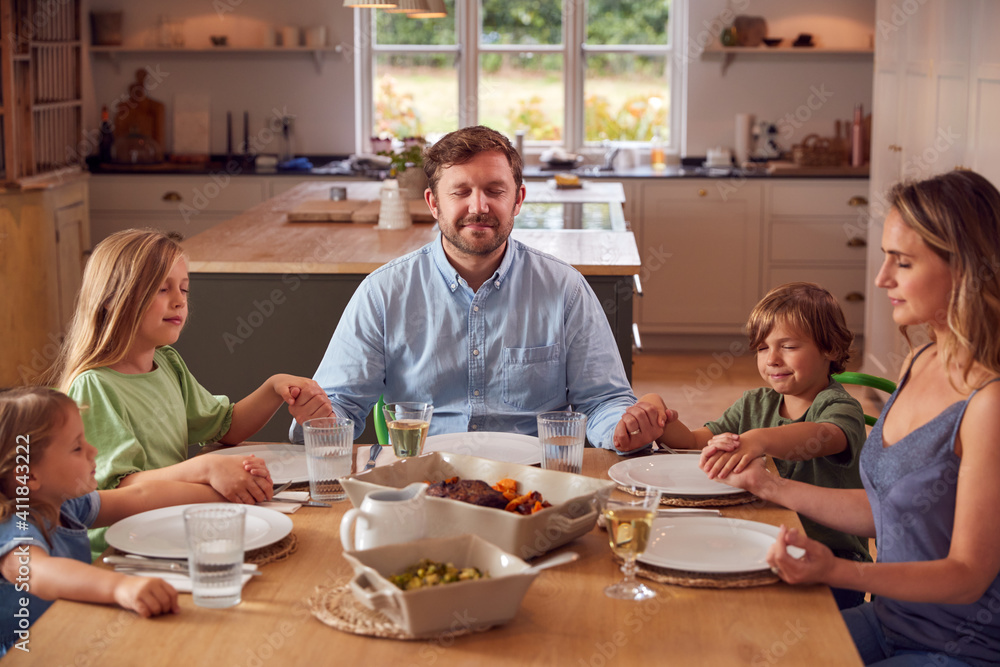 Family Sitting Around Table At Home Joining Hands And Saying Grace Before Meal Family Sitting Around Table At Home Joining Hands And Saying Grace Before Meal