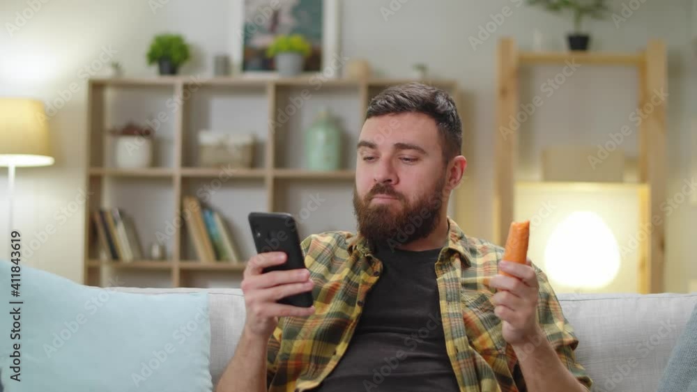Young handsome caucasian male eating carrot at home in living room sitting on sofa with phone Young handsome caucasian male eating carrot at home in living room sitting on sofa with phone