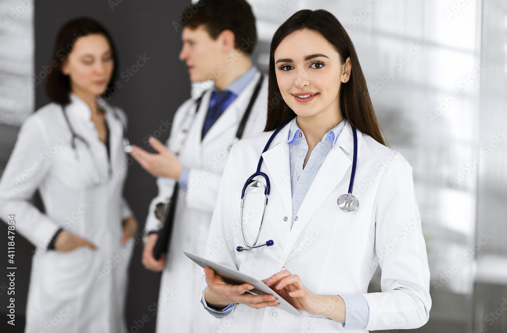 Smiling intelligent woman-doctor is holding a tablet computer in her hands, while she is standing together with her colleagues in a clinic. Physicians at work. Perfect medical service in a hospital Smiling intelligent woman-doctor is holding a tablet computer in her hands, while she is standing together with her colleagues in a clinic. Physicians at work. Perfect medical service in a hospital