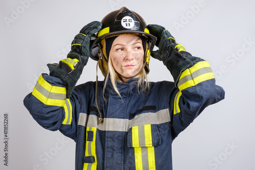 young brave woman in uniform of firefighter puts hardhat on her head and looking away on gray background