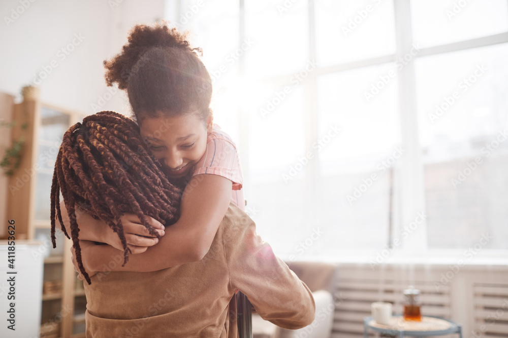 Minimal waist up portrait of happy African-American girl embracing mother in cozy home interior lit by sunlight, copy space - obrazy, fototapety, plakaty 