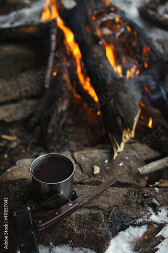 Metal mug with tea and a knife lying on a stone on the background of a burning fire , close-up.