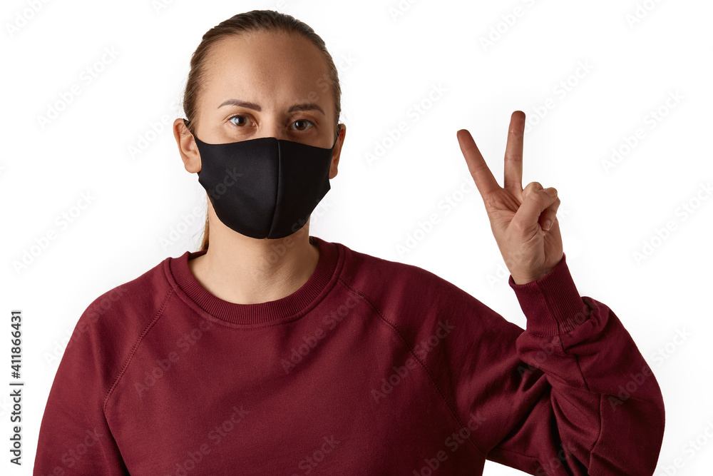 Peace. Young european woman with brown eyes wears face mask and burgundy sweatshirt, stands straight, shows victory gesture, number two, isolated over white background. Peace. Young european woman with brown eyes wears face mask and burgundy sweatshirt, stands straight, shows victory gesture, number two, isolated over white background.