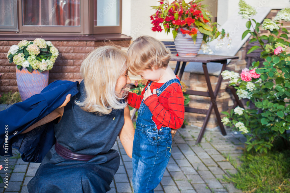 A child and an older woman hug and happily chat in the flower yard in front of the cottage. Grandmother greets her grandson. Joint time of the elder and the younger. Generational love. A child and an older woman hug and happily chat in the flower yard in front of the cottage. Grandmother greets her grandson. Joint time of the elder and the younger. Generational love.