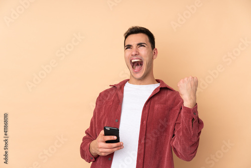 Young caucasian handsome man isolated on beige background with phone in victory position