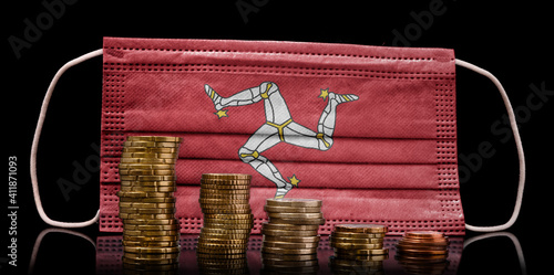 Medical mask with the flag of Isle of Man behind some shrinking stacks of various coins.(series) photo