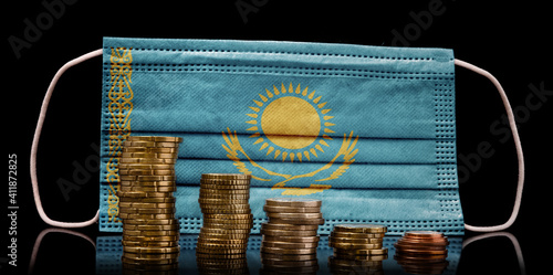 Medical mask with the flag of Kazakhstan behind some shrinking stacks of various coins.(series) photo