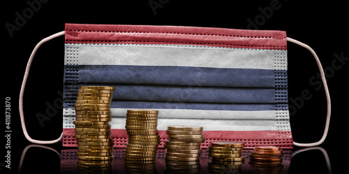 Medical mask with the flag of Thailand behind some shrinking stacks of various coins.(series) photo