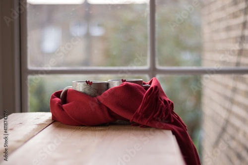 Love and Valentine's Day Mood - romantic minimalism concept. Two mugs with red hearts tied one red scarf are on wooden table. Background is window in simple room. Creative Still life Composition.