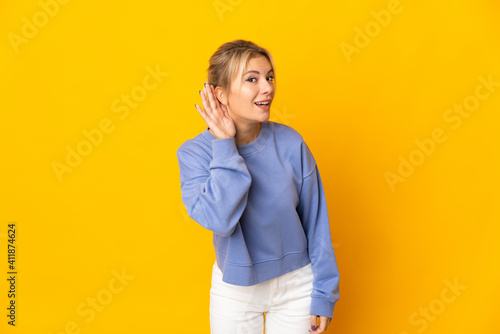Young Russian woman isolated on yellow background listening to something by putting hand on the ear