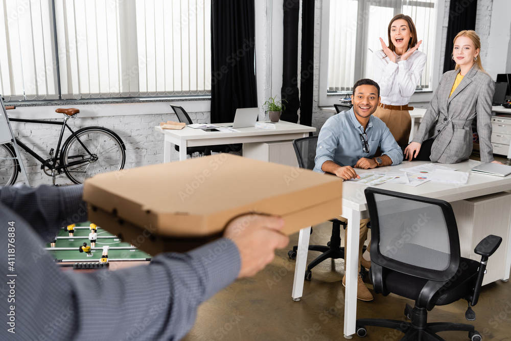 Obraz premium Cheerful multiethnic businesspeople looking at delivery man with pizza boxes on blurred foreground