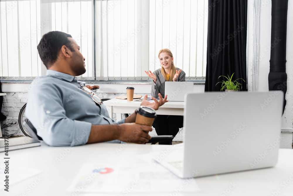 Smiling businesswoman talking with indian colleague with coffee to go near laptop in office Smiling businesswoman talking with indian colleague with coffee to go near laptop in office