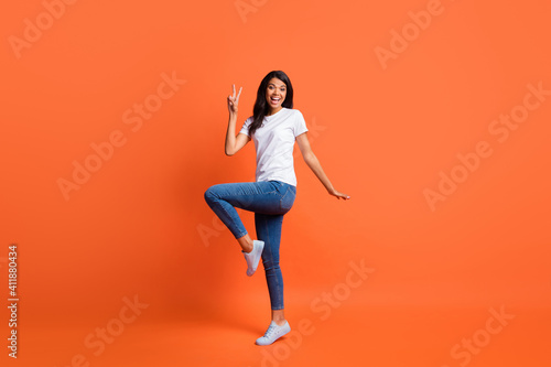Full length photo of lady show v-sign open mouth wear white t-shirt jeans footwear isolated orange color background
