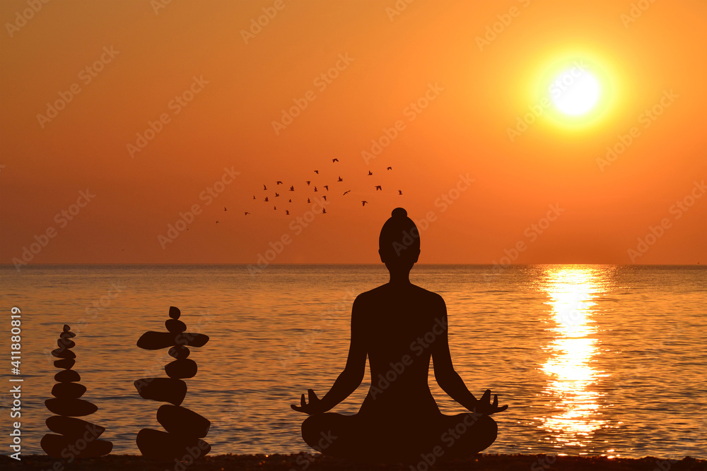 Silhouette of yogi in lotus position and a piles of stones at sea shore at sunrise