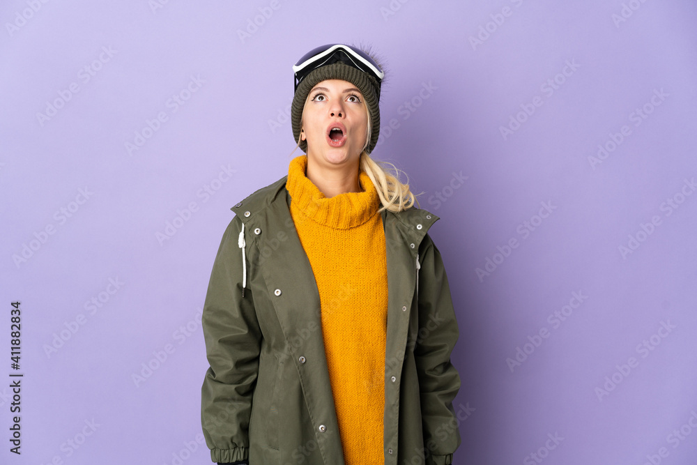 Skier Russian girl with snowboarding glasses isolated on purple background looking up and with surprised expression Skier Russian girl with snowboarding glasses isolated on purple background looking up and with surprised expression