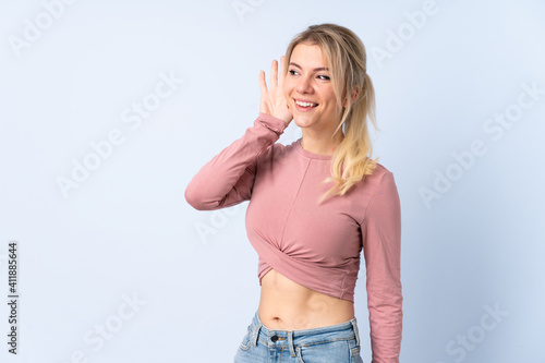 Blonde woman over isolated blue background listening to something by putting hand on the ear