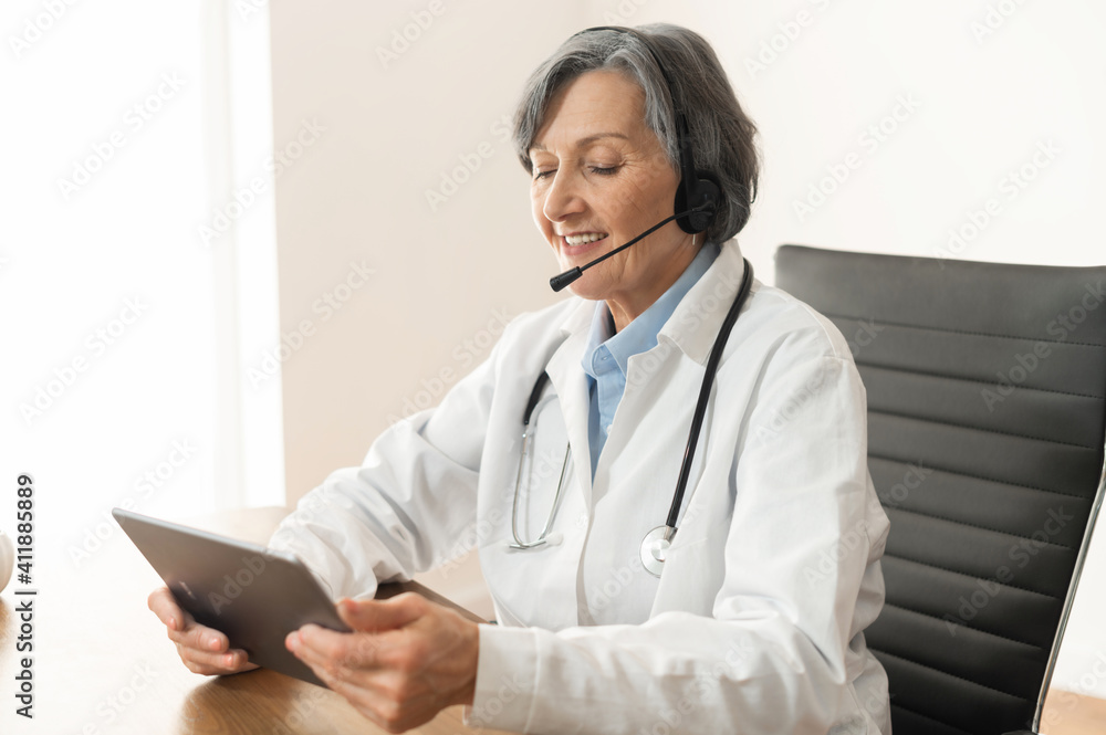 Senior old female doctor in a lab coat sitting at the desk in her office with headset, consulting a patient on the insurance and treatment plan on a video call, telehealth or telemedicine concept