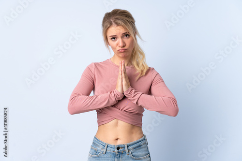 Blonde woman over isolated blue background keeps palm together. Person asks for something