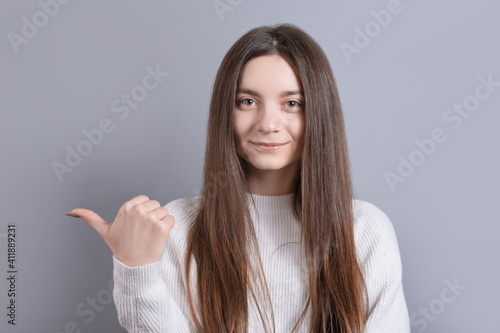 Young beautiful woman portrait with dark hair wearing white sweater smiling with happy face looking and pointing to the side with thumb up.Copy space.