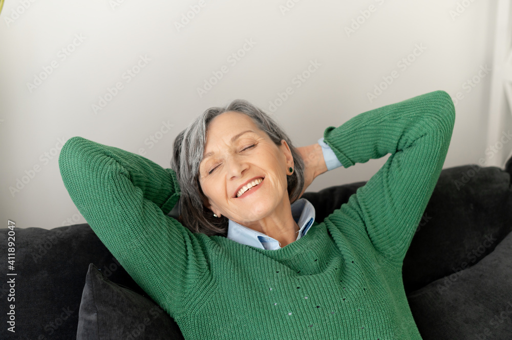 Smiling senior mature lady is resting on the couch or sofa with eyes closed, placed her hands behind the head, relaxing after a long working day, having a great mood Smiling senior mature lady is resting on the couch or sofa with eyes closed, placed her hands behind the head, relaxing after a long working day, having a great mood