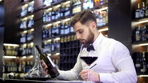Wine cellar. Handsome sommelier tasting red wine and making notes at degustation card
