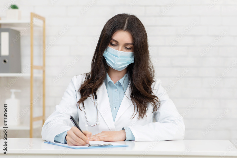 Young serious female doctor in white coat and protective mask writes prescription to clinic patient Young serious female doctor in white coat and protective mask writes prescription to clinic patient