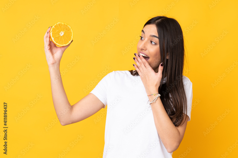 Young caucasian woman holding an orange isolated on yellow background with surprise and shocked facial expression
