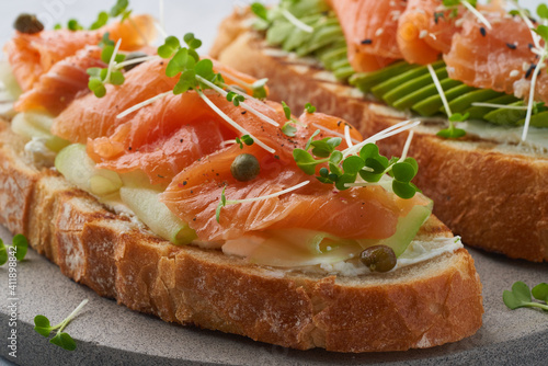 Two open sandwich, toast with salmon, cream cheese, avocado, cucumber slices on white concrete table. Morning healthy breakfast with fish and cup of drink, macro, close up