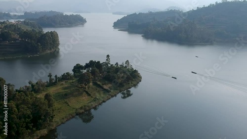 Three small boats, mokoros, transporting people on the calm waters of the lake, you can see the wake of the boats trailing behind on the water. green hills with trees all around the lake. Aerial Video photo