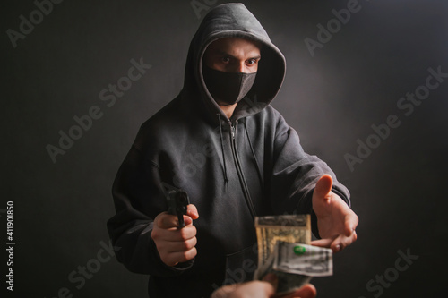 A gangster in a hood and a mask holds a weapon in his hands on a dark background and takes money from the victim. The bandit aims his pistol and demands dollars. Photo in the studio.