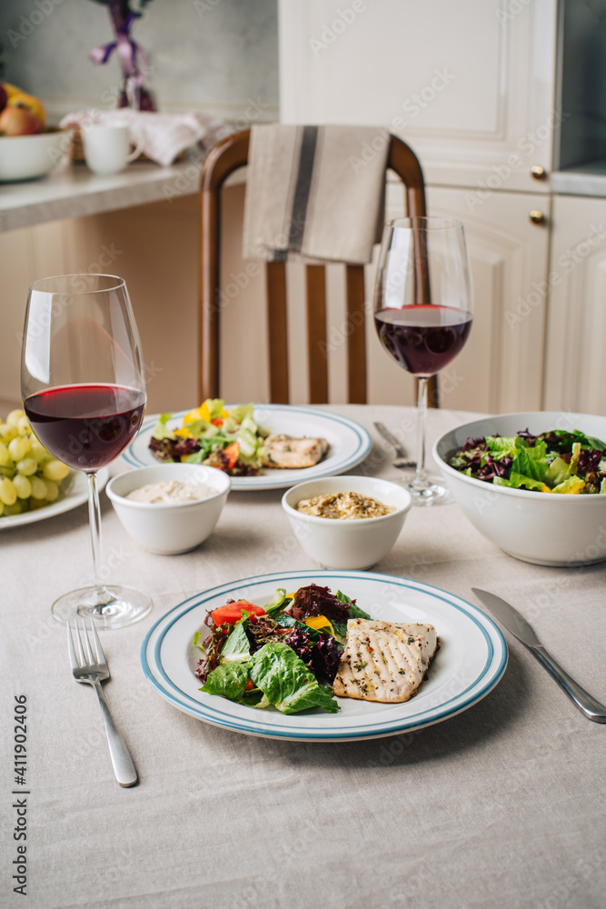 White fish steak with vegetable salad, sauce, hummus, grapes and glasses of wine. Served for two in the kitchen