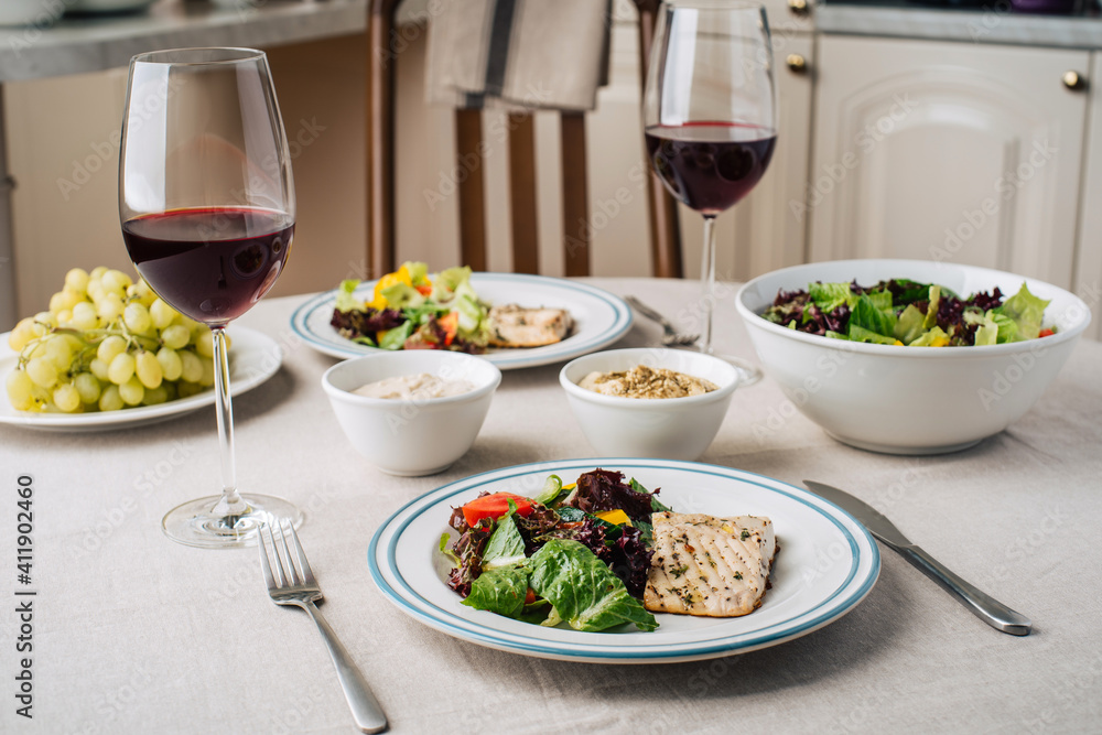 White fish steak with vegetable salad, sauce, hummus, grapes and glasses of wine. Served for two in the kitchen White fish steak with vegetable salad, sauce, hummus, grapes and glasses of wine. Served for two in the kitchen