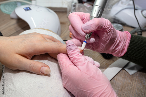 Removing the old hybrid nail using an electric nail grinder, the beautician has pink latex gloves.