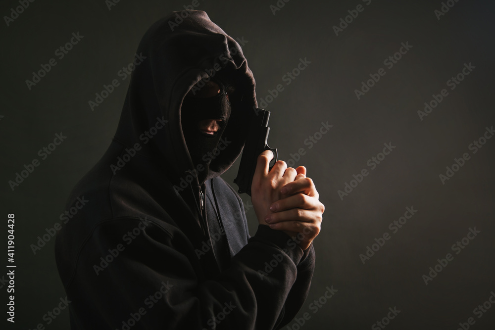 A gangster in a hood, a mask, holds a weapon in his hands on a dark background. The bandit aims with a pistol. A gangster in a hood, a mask, holds a weapon in his hands on a dark background. The bandit aims with a pistol.