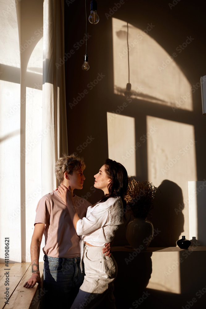 Side view of loving couple tenderly hugging while standing near windowsill in room lit by sunlight and looking at each other Side view of loving couple tenderly hugging while standing near windowsill in room lit by sunlight and looking at each other