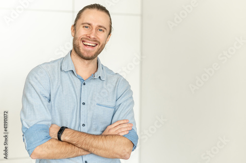 Handsome hipster guy stands with his arms crossed, looks at the camera and laughs. Portrait of cheerful young man wearing smart casual shirt on the white background, copy space