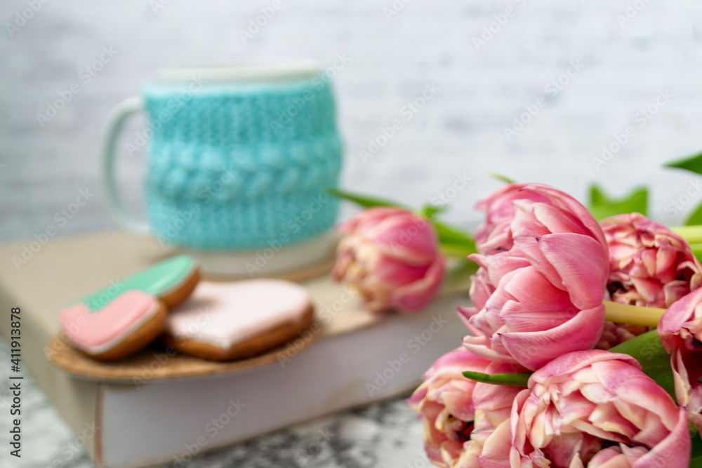 A white cup of tea in a knitted blue case stands on a book and pink flowers, tulips and gingerbread on a texture table. Spring concept. A white cup of tea in a knitted blue case stands on a book and pink flowers, tulips and gingerbread on a texture table. Spring concept.