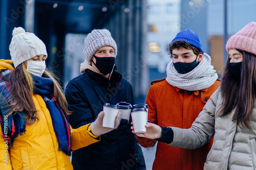 Group of four young people in facial protective masks meeting outdoor in winter day. New normal lifestyle concept with happy guys and girls.